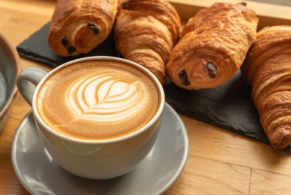 Ein Cappuccino mit Blatt-Latte-Art neben frisch gebackenen Croissants und Pain au chocolat auf warmem Holz.